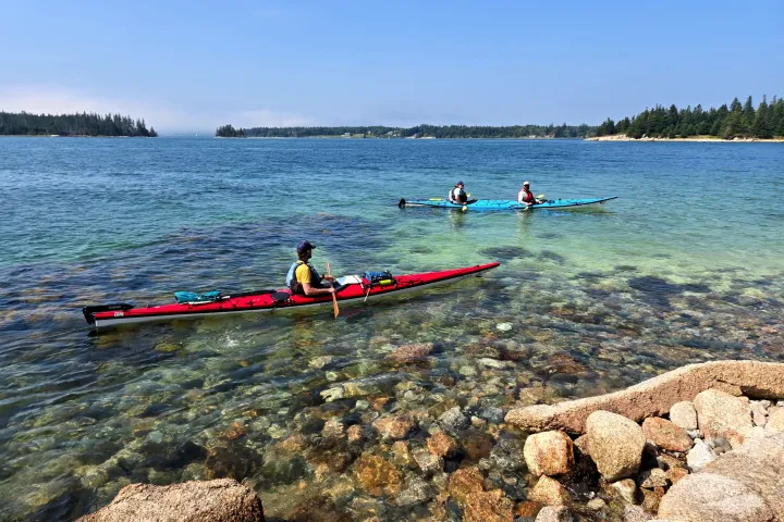 Three kayakers on clear water near a rocky shoreline with distant trees.