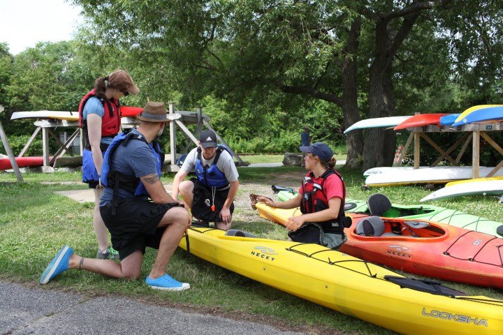 Four people in life vests talking near colorful kayaks on a grassy area.