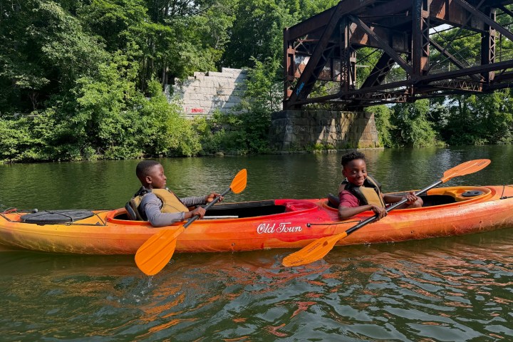 Two children kayaking on a river under a metal bridge.