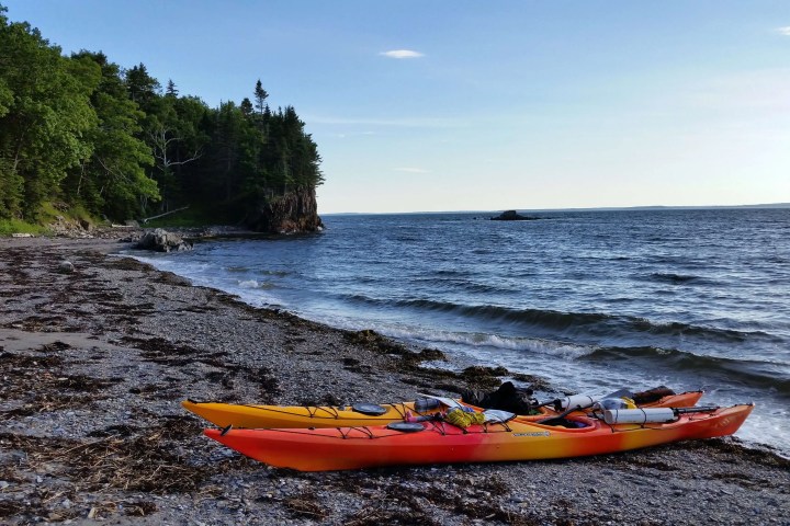 Two kayaks on a rocky beach beside a forested coastline with ocean waves.