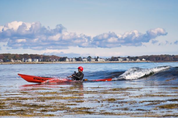 Person in red kayak with helmet paddling near a wave in the ocean.