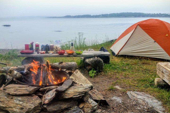 Campfire near a tent by a lake, with a picnic table of food in the background.