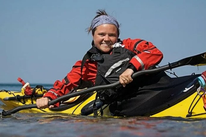 Person in red jacket kayaking on clear blue water, smiling, with sky in background.