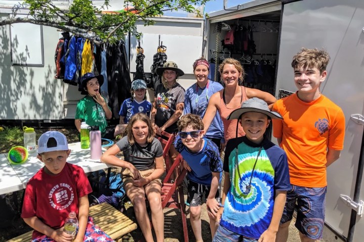 Group of kids and adults posing outdoors near a trailer with gear.