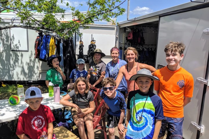 Group of kids and adults posing outdoors near a trailer with gear.