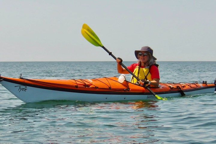 Person kayaking on calm sea in orange kayak, wearing a yellow life vest, red shirt, and wide-brim hat.