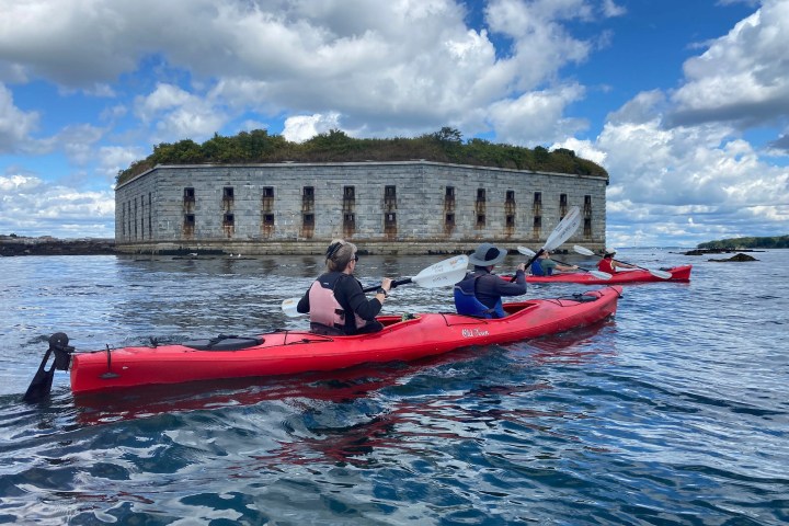People kayaking in red kayaks near a stone fort under a partly cloudy sky.