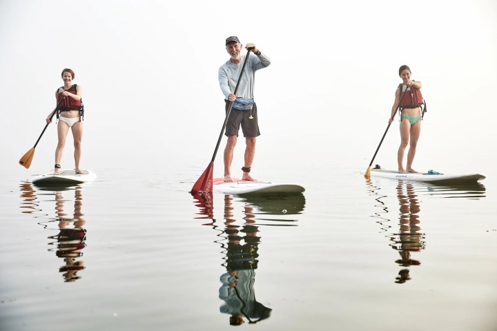 Three people paddleboarding on calm water, wearing life jackets, with reflections visible on the water surface.