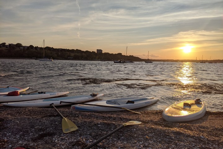 Paddleboards and paddles on a beach at sunset with boats on the water.