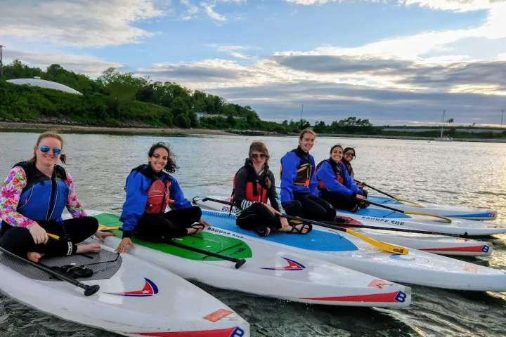 Six people in life vests sitting on paddleboards in a river, with trees and a cloudy sky in the background.