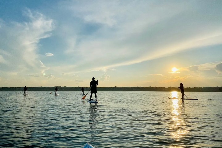 People paddleboarding on a calm lake at sunset with a clear sky.