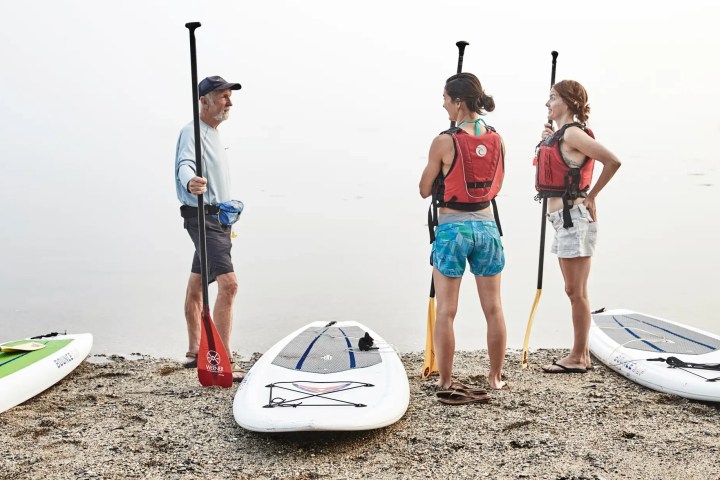 Three people with paddles beside paddleboards on a misty beach.