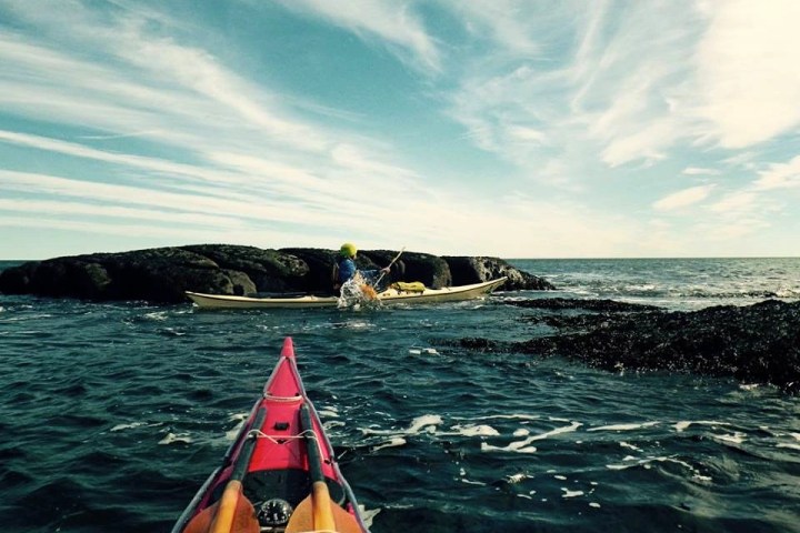 Two kayakers on the ocean near a rocky formation under a partly cloudy sky.