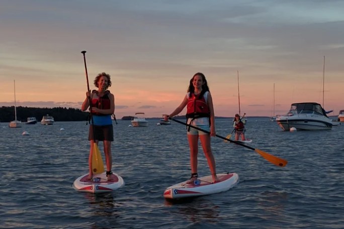 Two people paddleboarding on a calm sea at sunset with boats in the background.