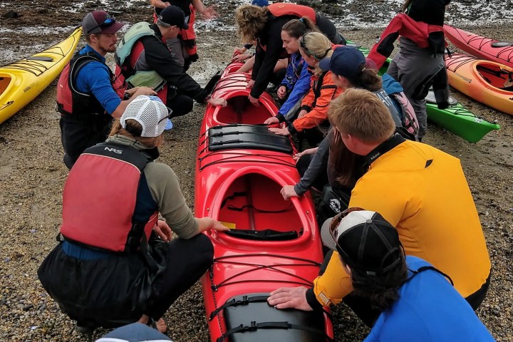 Group gathered on shore around red kayak, listening to instructor.