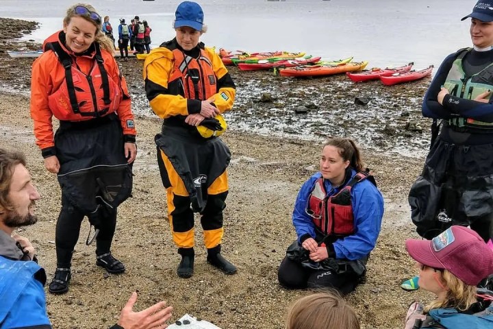 Group in kayaks and gear gathered on beach with map, listening to guide.