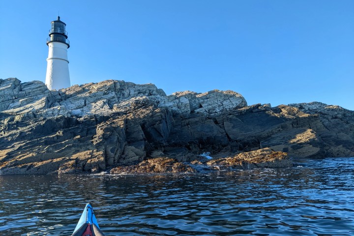View of a lighthouse on rocky cliffs from a kayak in the water under a clear blue sky.