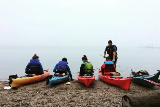 Four people by kayaks on a foggy beach, preparing to enter the water.
