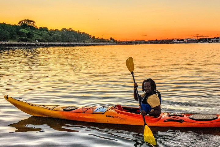 Person kayaking on a calm river at sunset with trees and buildings in the background.