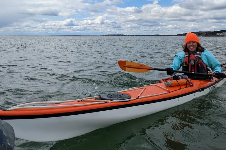 Person kayaking on ocean in orange kayak, wearing a bright orange hat and teal jacket under cloudy sky.