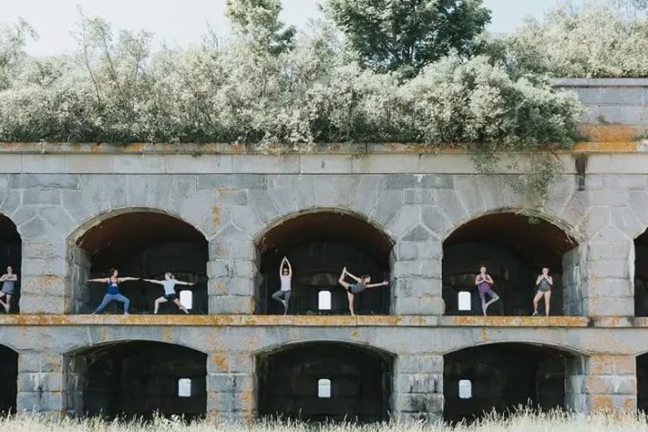 People doing yoga poses in arches of a stone structure with greenery on top.