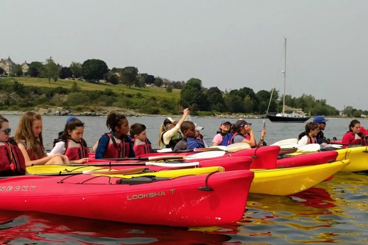 Group of children in red and yellow kayaks on a lake, wearing life jackets.