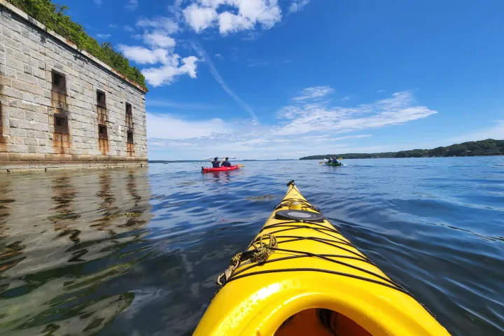 View from a yellow kayak near stone Fort and other kayakers on calm blue water under clear sky.