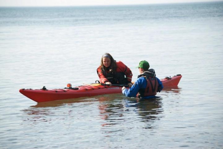 Two people in water, one in a red kayak, the other standing, in outdoor gear.