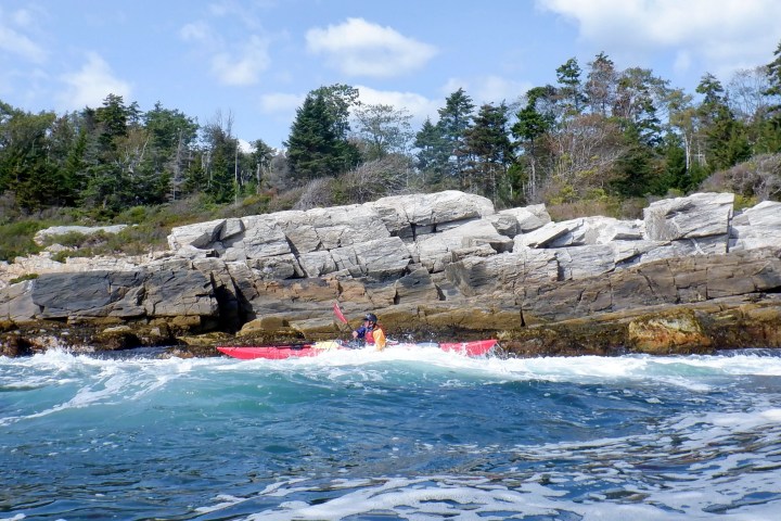 A kayaker paddles near rocky shoreline with trees under a blue sky.