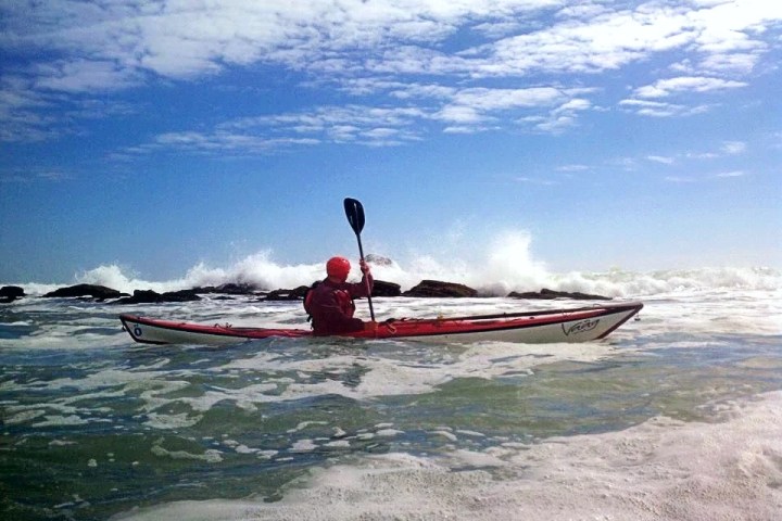 Kayaker in red gear paddling through ocean waves under a cloudy blue sky.