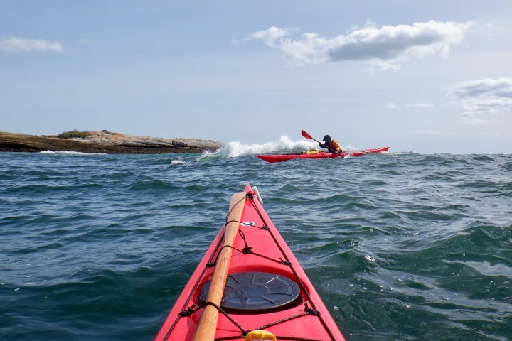 Two kayakers in red kayaks paddling on a wavy sea near a rocky shore.