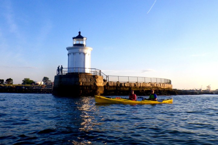 Two people kayaking near a lighthouse on a sunny day.