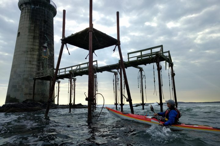 Sea kayak below a pier and lighthouse which seem to rise directly from the water - Ram Island Ledge Light