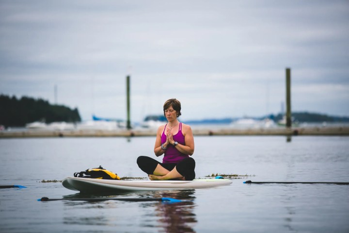 Person meditating on a paddleboard on calm water with a cloudy sky.