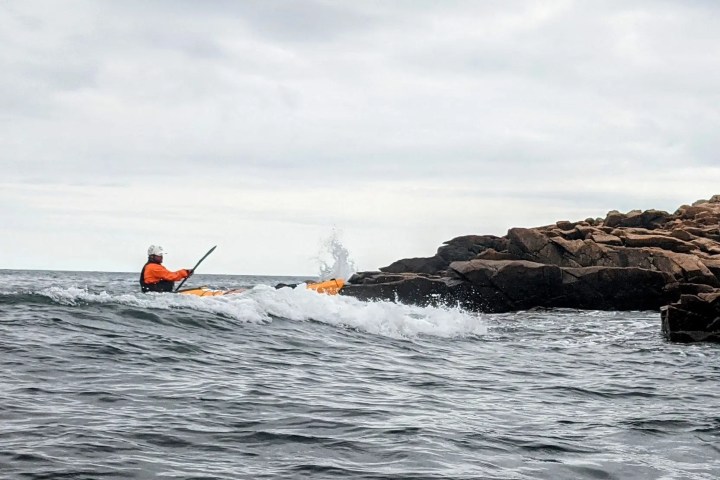Kayaker in orange jacket paddles near rocky shore on choppy sea.