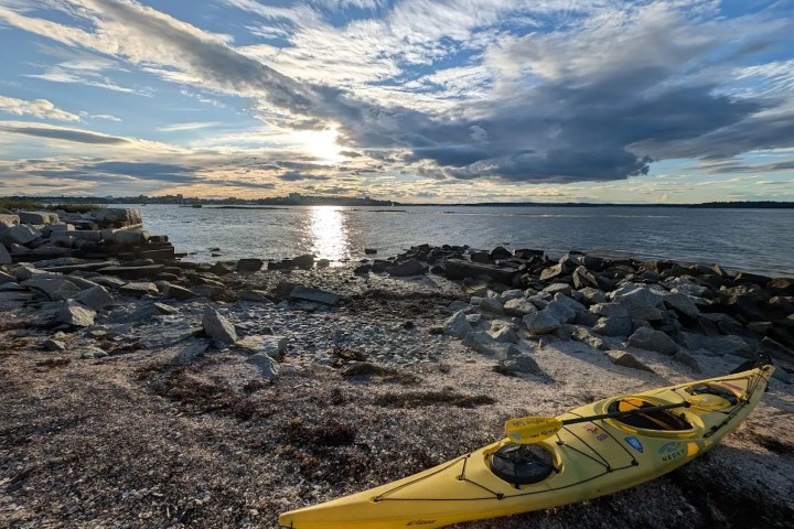 Yellow kayak on rocky shore with ocean and cloudy sky at sunset.