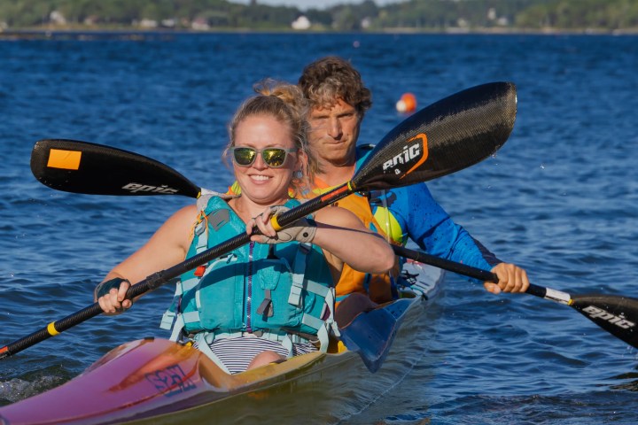 Two people kayaking in a tandem kayak on a sunny day with water and trees in the background.