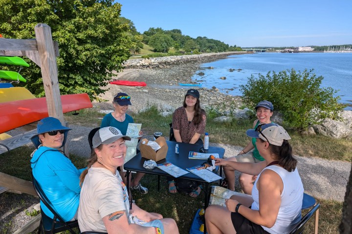 Group of six people smiling and sitting at a table by a lake with colorful kayaks in the background.
