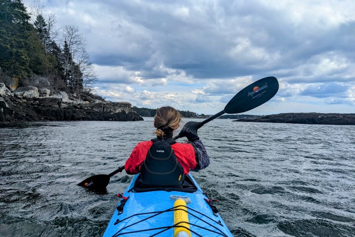 Person kayaking on a lake with rocky shoreline and cloudy sky.