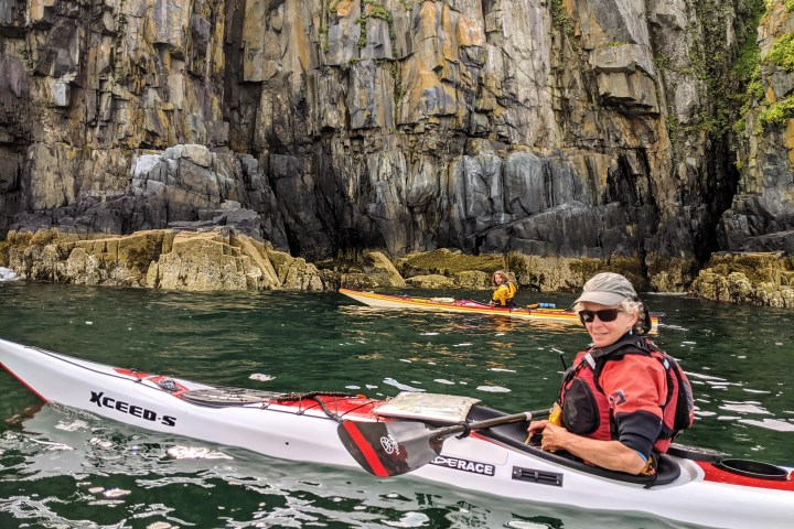Two kayakers in colorful attire paddle near rocky coastal cliffs.