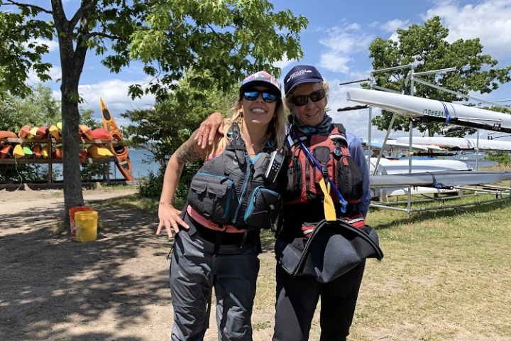 Two people in life jackets smiling by a lake with kayaks and trees nearby.