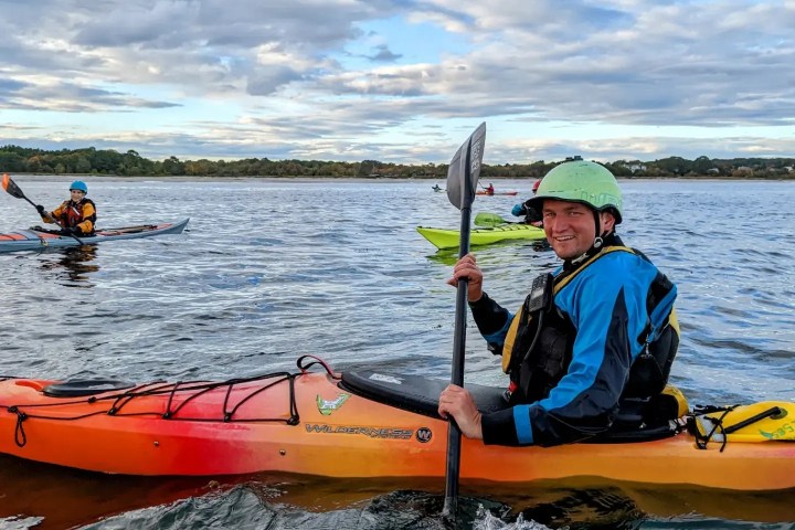 Person kayaking on a lake with a helmet and paddle, smiling at the camera.