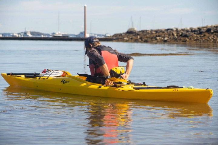 Person in a red life vest adjusts gear in a yellow kayak on calm water by a rocky shore.