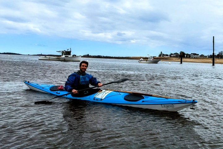 Person kayaking on calm water with boats and shoreline in the background.