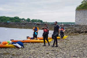 A group of people and their kayaks standing on a cobble and boulder beach