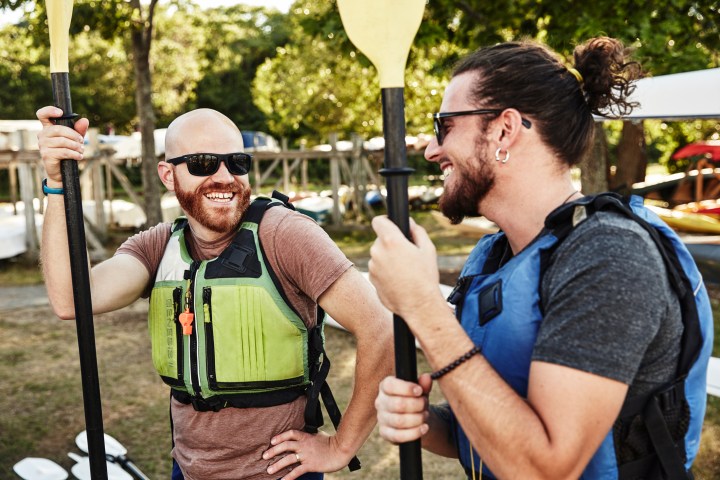 Two men in life vests smiling and holding paddles outdoors near kayaks.