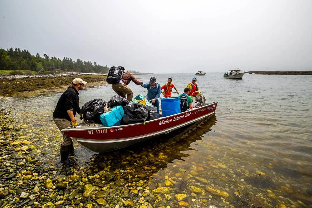 Group unloads trash bags and barrels from a boat onto a rocky shore in a foggy setting.
