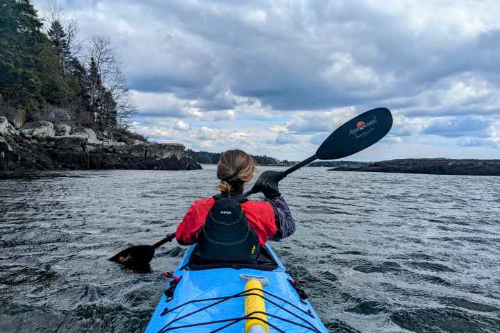 Person kayaking on a lake with rocky shoreline and cloudy sky.