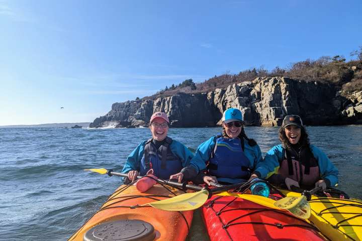 Three kayakers in colorful gear near a rocky cliff with a clear blue sky.