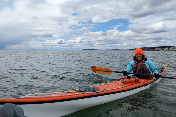 Person in orange kayak wearing an orange beanie, paddling on open water under a cloudy sky.